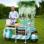 A man in a white shirt presenting a Pampelle bottle at a stylish stand surrounded by greenery, decorated with plants and an LED Pampelle sign