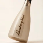 A minimalist shot of a tilted, gold, polygonal bottle of Lamborghini Icona Oro sparkling wine with elegant embossed lettering on a light background.
