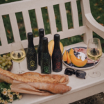 Four small bottles of Lamborghini Brut Mini Fluo sparkling wine with neon patterns on a white bench during a picnic. Next to them are glasses, baguettes, and a plate with melon and snacks.