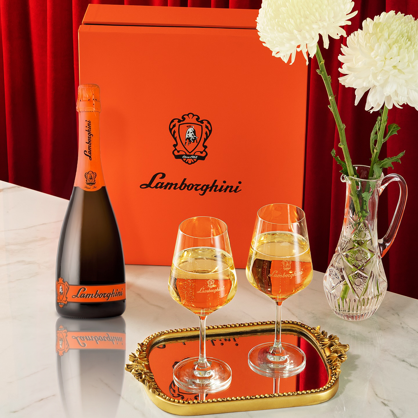 Luxury arrangement on a marble countertop with a red curtain in the background: a bottle of Lamborghini Demi Sec Prosecco DOCG, a large orange gift box, two glasses of wine on a gold mirrored tray, and a crystal vase with white chrysanthemums