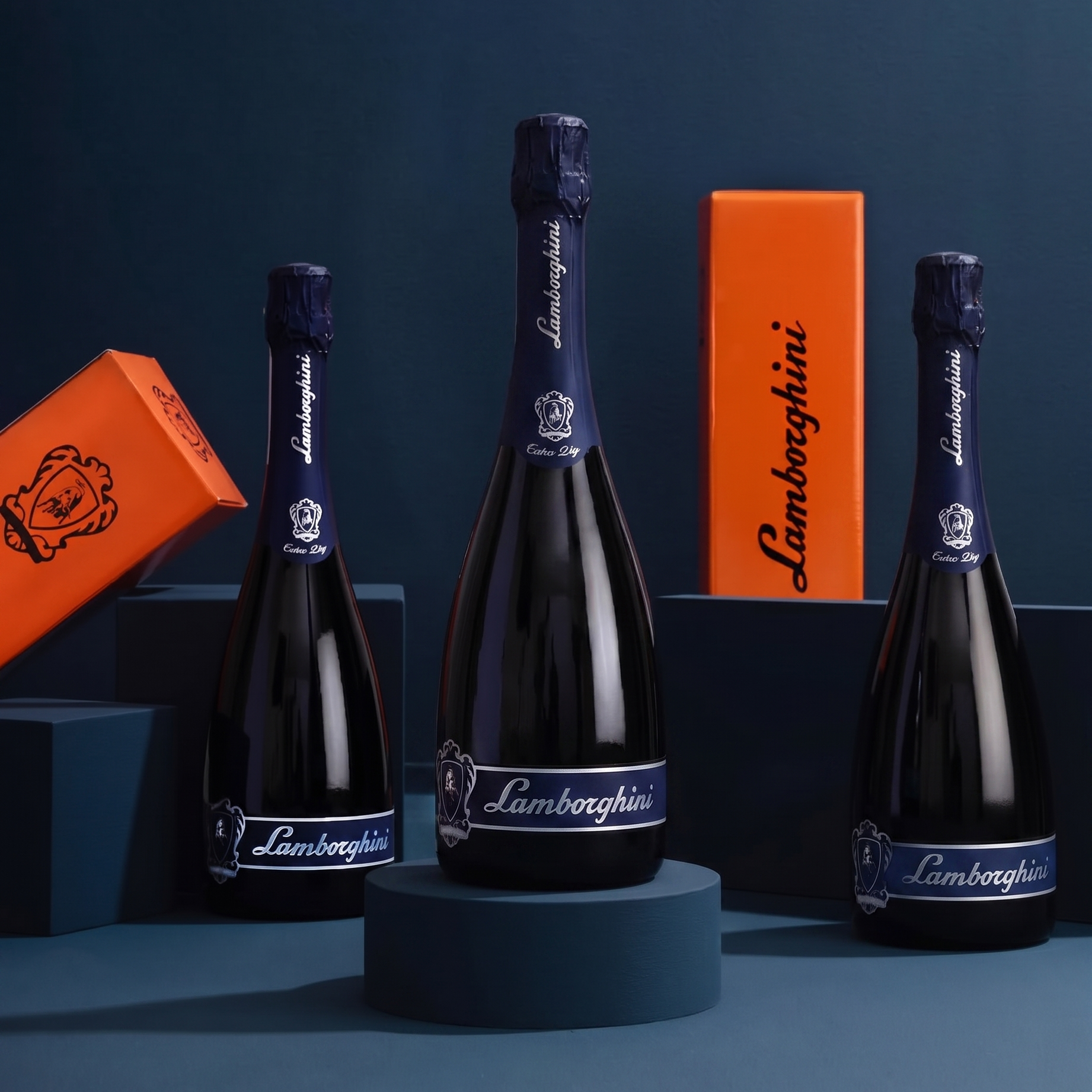 Three elegant black bottles of Lamborghini Extra Dry Prosecco with blue details, presented on geometric navy blue pedestals accompanied by two vertical orange Lamborghini gift boxes.