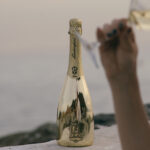 A gold bottle of Lamborghini Extra Dry Gold standing on a wall against the backdrop of a calm sea at dusk. In the blurred foreground, a hand holding a wine glass.
