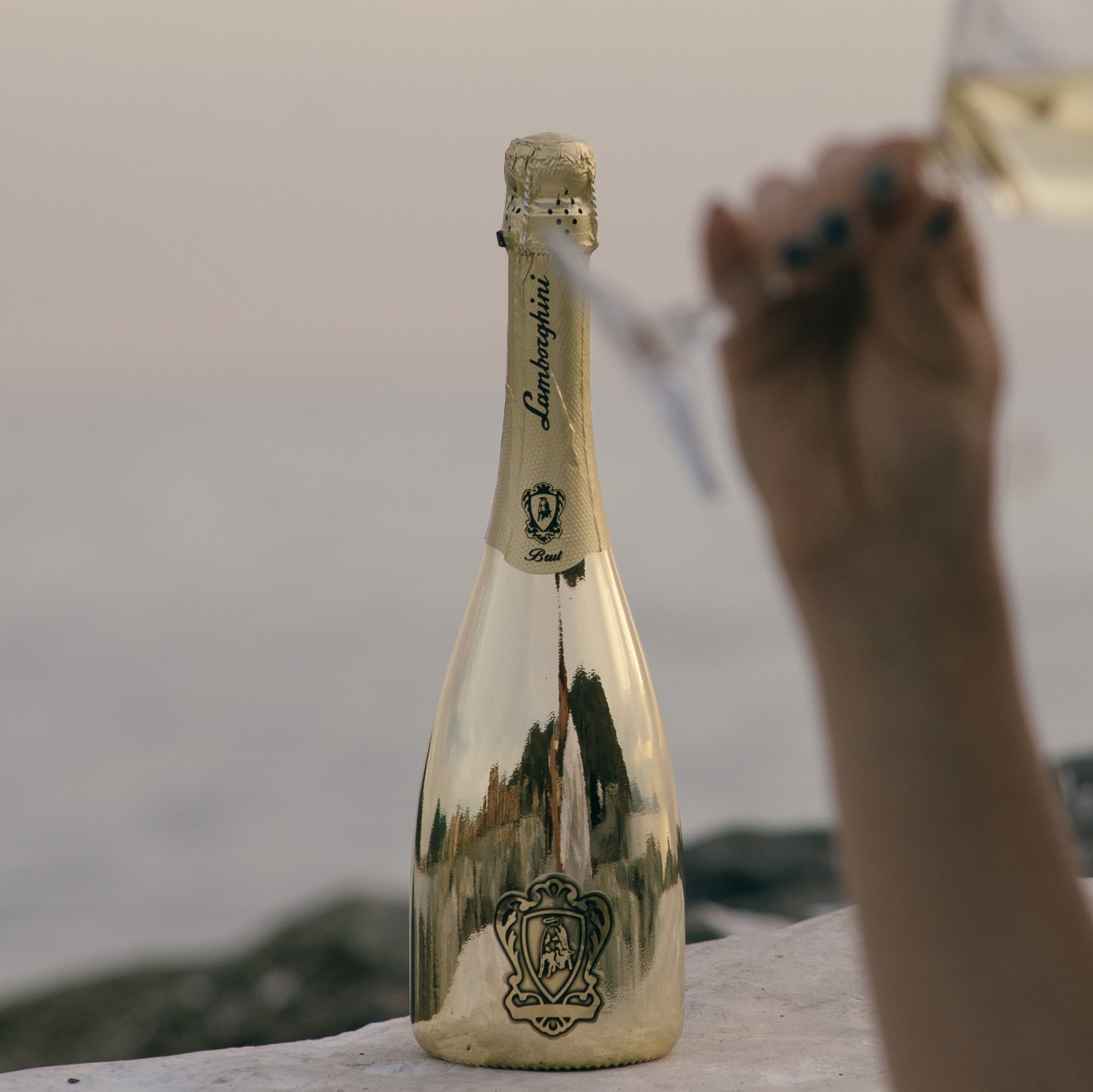 A gold bottle of Lamborghini Extra Dry Gold standing on a wall against the backdrop of a calm sea at dusk. In the blurred foreground, a hand holding a wine glass.