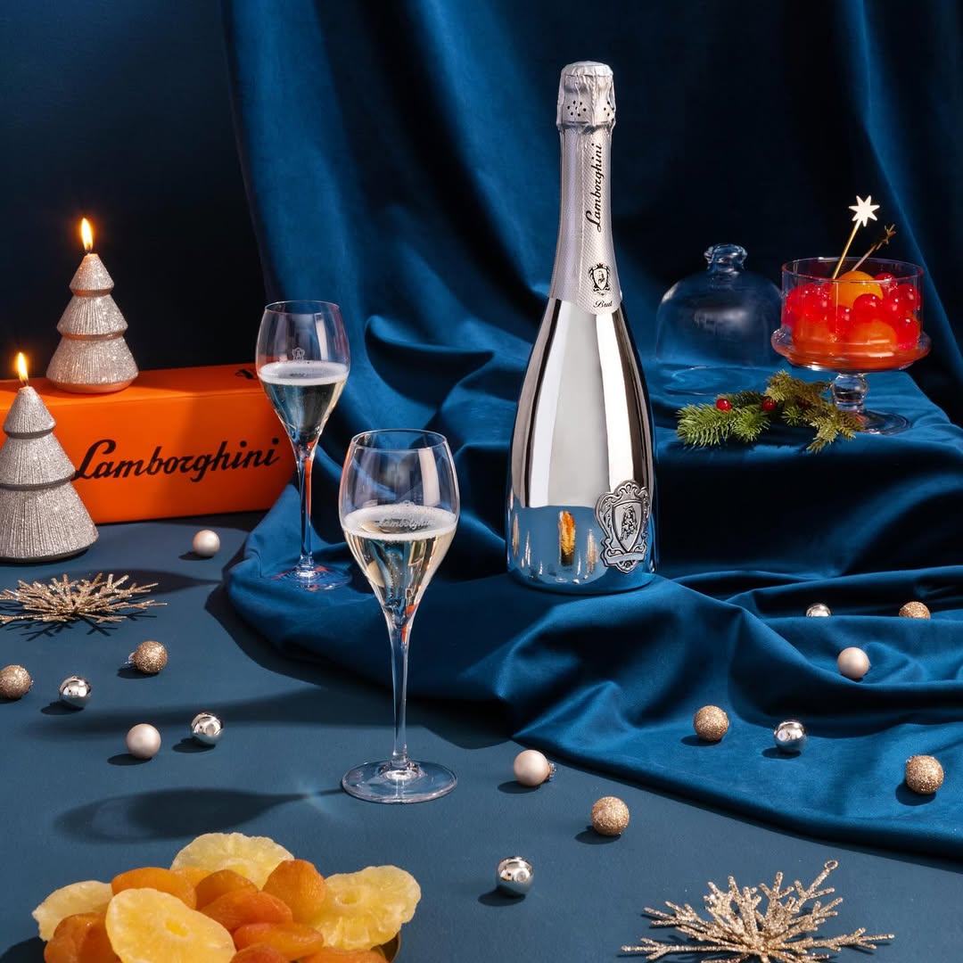 A silver bottle of Lamborghini Extra Dry Silver wine and two glasses against an elegant dark blue velvet background. Festive decorations around: silver baubles, tree-shaped candles, candied fruits, and an orange box.