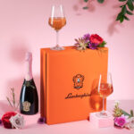 A composition featuring a bottle of Lamborghini Metodo Classico Rosé wine, two filled glasses, and a large orange gift box with the brand's logo. The setting is decorated with flowers on a pink background.