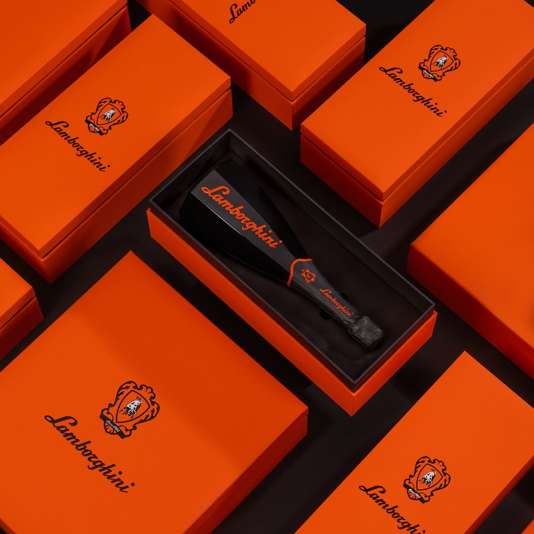 A composition of many closed orange Lamborghini gift boxes arranged on a black background. In the center, one open box displays a black and orange bottle of Ottagonale Brut wine.