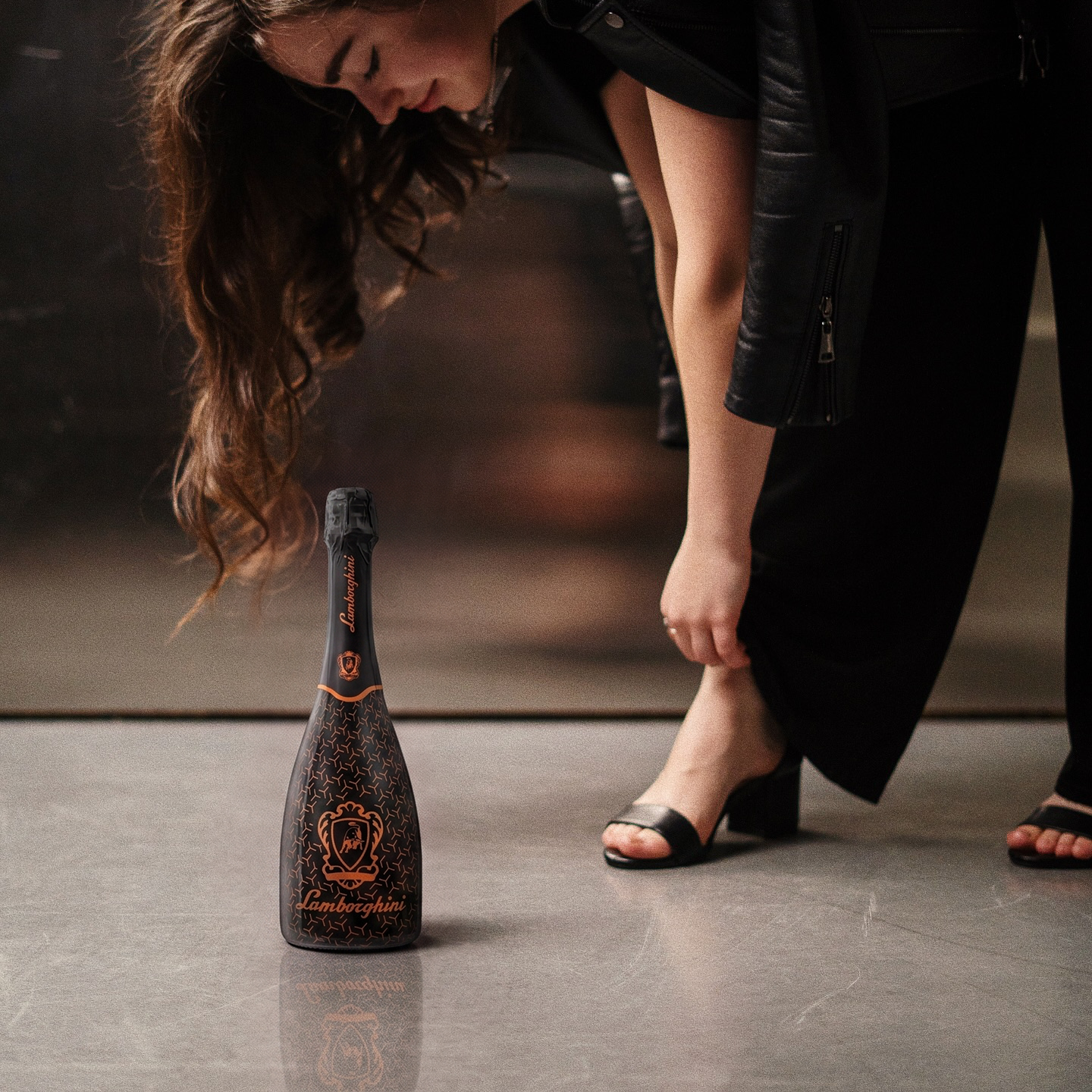 A woman in a black leather jacket and block-heel sandals leans down on a concrete floor near a bottle of Lamborghini sparkling wine with a bronze/orange pattern. Light-reflecting metal background.