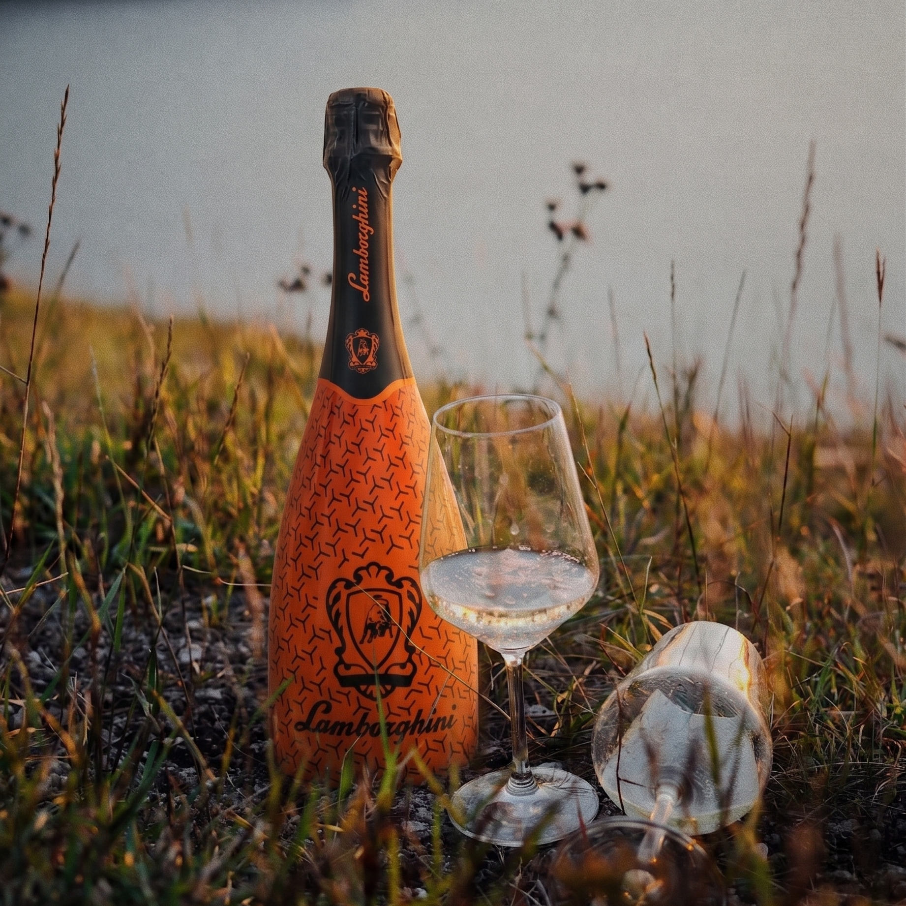 An orange bottle of Lamborghini Demi Sec wine in geometric patterns, standing in tall grass at sunset next to a filled glass and a second, overturned one. The background is water and a summer landscape