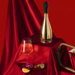 A gold, mirrored bottle of non-alcoholic Lamborghini 0% Gold wine placed on an elegant, draped red velvet. Next to it is a filled glass, a gold stopper, and an orange slice.