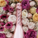 A pink, metallic bottle of non-alcoholic Lamborghini 0% Rosé sparkling wine lying on a dense, romantic background of fresh roses in shades of fuchsia, light pink, and white.
