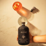Flatlay shot. A hand holding a black Mezcal Don Ramón Salmiana Joven bottle just above a glass with ice. Next to it is a finished orange cocktail with a lemon peel and the jaguar stopper lying beside it.