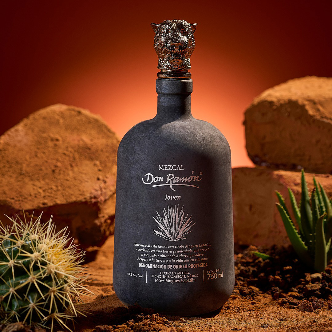 A black, matte bottle of Mezcal Don Ramón Joven with a decorative jaguar head stopper, standing on red earth among desert rocks, agave, and a cactus against an orange background.