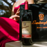 A bottle of Lamborghini Centanni Rosso Umbria red wine standing in the sun outdoors, next to an elegant black Lamborghini gift box. In the background, a woman in a red dress under a pergola is visible.