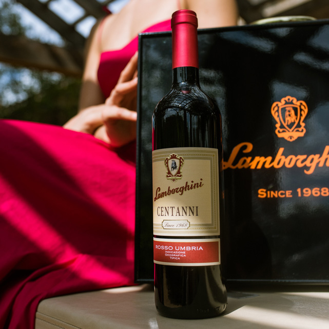 A bottle of Lamborghini Centanni Rosso Umbria red wine standing in the sun outdoors, next to an elegant black Lamborghini gift box. In the background, a woman in a red dress under a pergola is visible.