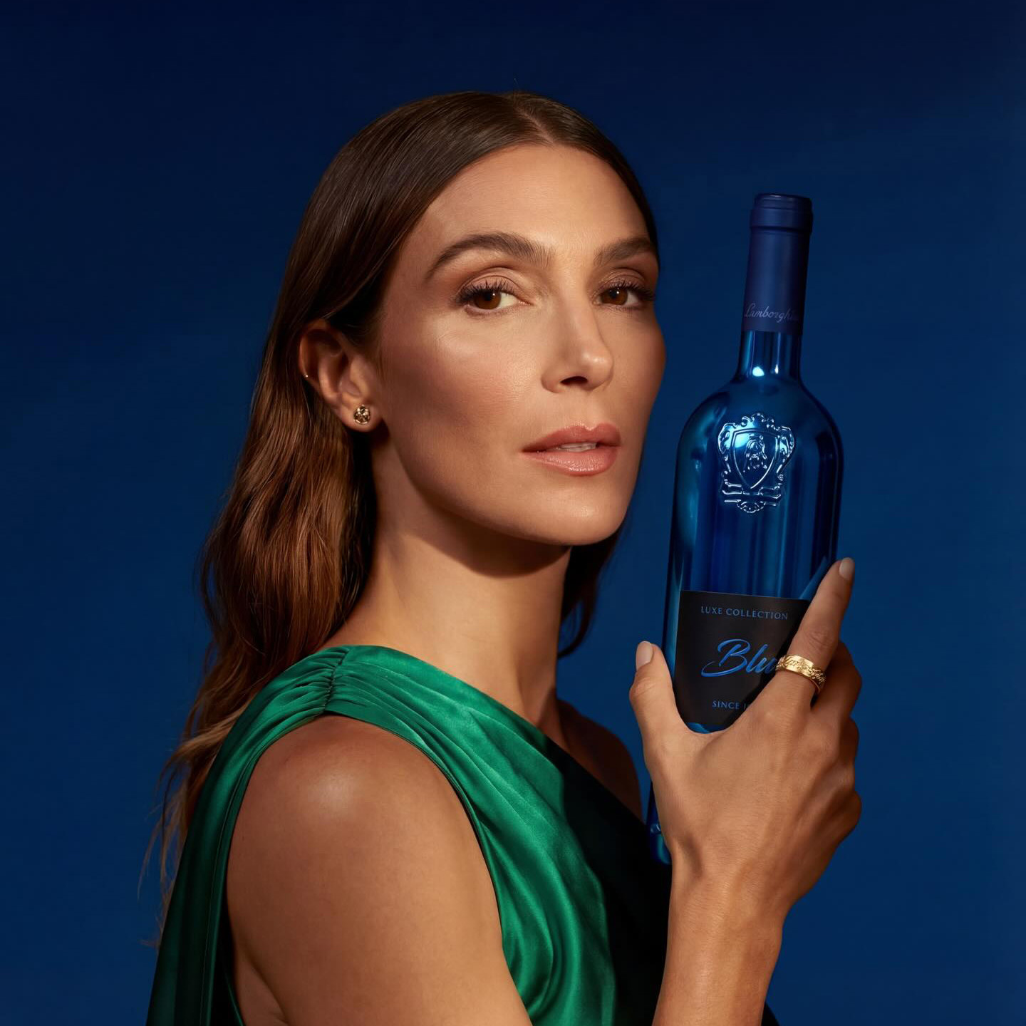 A woman of subtle beauty, wearing a green silk dress, presenting a metallic blue bottle of Lamborghini Luxe Blu wine against a deep, dark blue background.