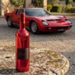 A shiny red Lamborghini Luxe Rosso wine bottle standing on a rustic stone table. In the background, a classic red Lamborghini Miura car is parked in the sun in front of an old Italian villa.