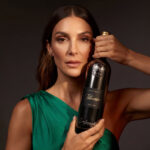 An elegant woman in an emerald green silk dress, holding a dark, metallic bottle of Lamborghini Luxe Titanio red wine against a warm, dark background.