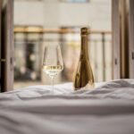 A gold bottle of Lamborghini Extra Dry and a glass of white wine resting on white bedding in a bedroom. In the background, an open window overlooking a balcony and historic architecture.
