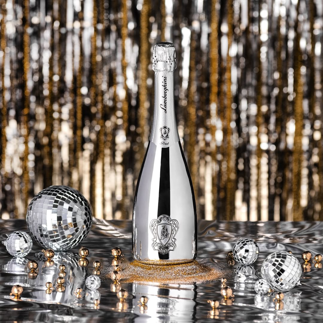 A silver, mirrored bottle of Lamborghini Extra Dry Silver surrounded by small disco balls, gold glitter, and pearls against a shiny silver and gold party curtain background.