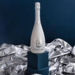 A phenomenal white bottle of Lamborghini Ice Brut wine displayed on elegant navy blue geometric pedestals, surrounded by wavy, shiny silver fabric.