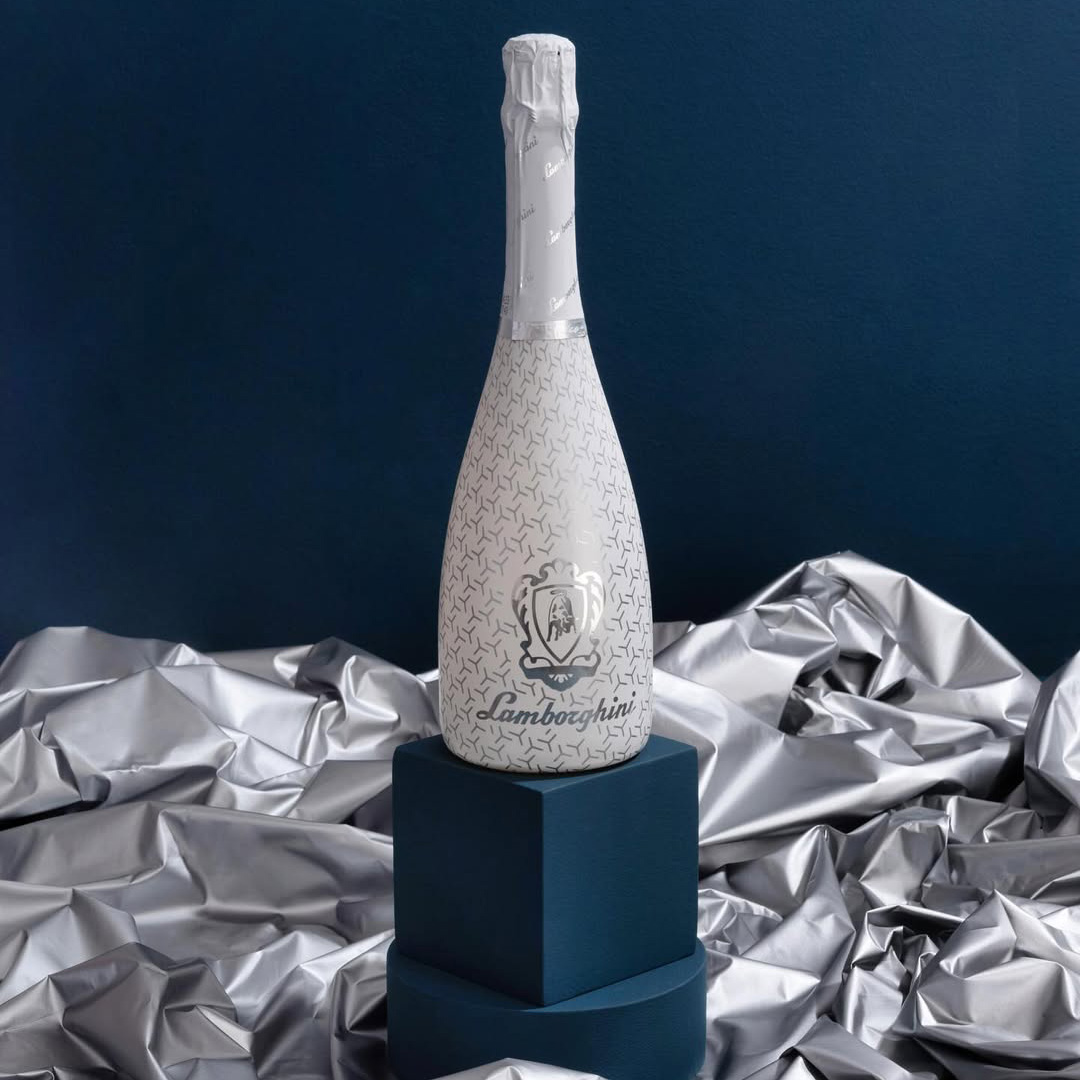A phenomenal white bottle of Lamborghini Ice Brut wine displayed on elegant navy blue geometric pedestals, surrounded by wavy, shiny silver fabric.