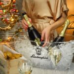A woman in a gold dress chilling a bottle of Lamborghini Metodo Classico Brut wine in a transparent ice bucket. Christmas lights in the background.