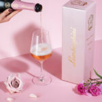 A hand pouring Lamborghini Metodo Classico Rosé sparkling wine into an elegant glass on a light pink background with flowers and a luxury box.