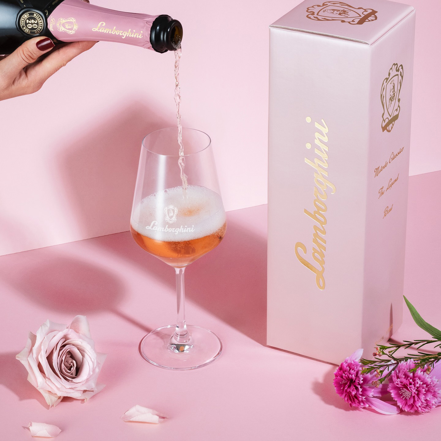 A hand pouring Lamborghini Metodo Classico Rosé sparkling wine into an elegant glass on a light pink background with flowers and a luxury box.