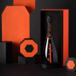 A black bottle of Lamborghini Ottagonale Brut wine with a unique octagonal shape and orange lettering, placed in an open orange and black box against a background of geometric shapes.