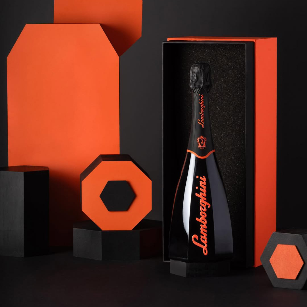 A black bottle of Lamborghini Ottagonale Brut wine with a unique octagonal shape and orange lettering, placed in an open orange and black box against a background of geometric shapes.