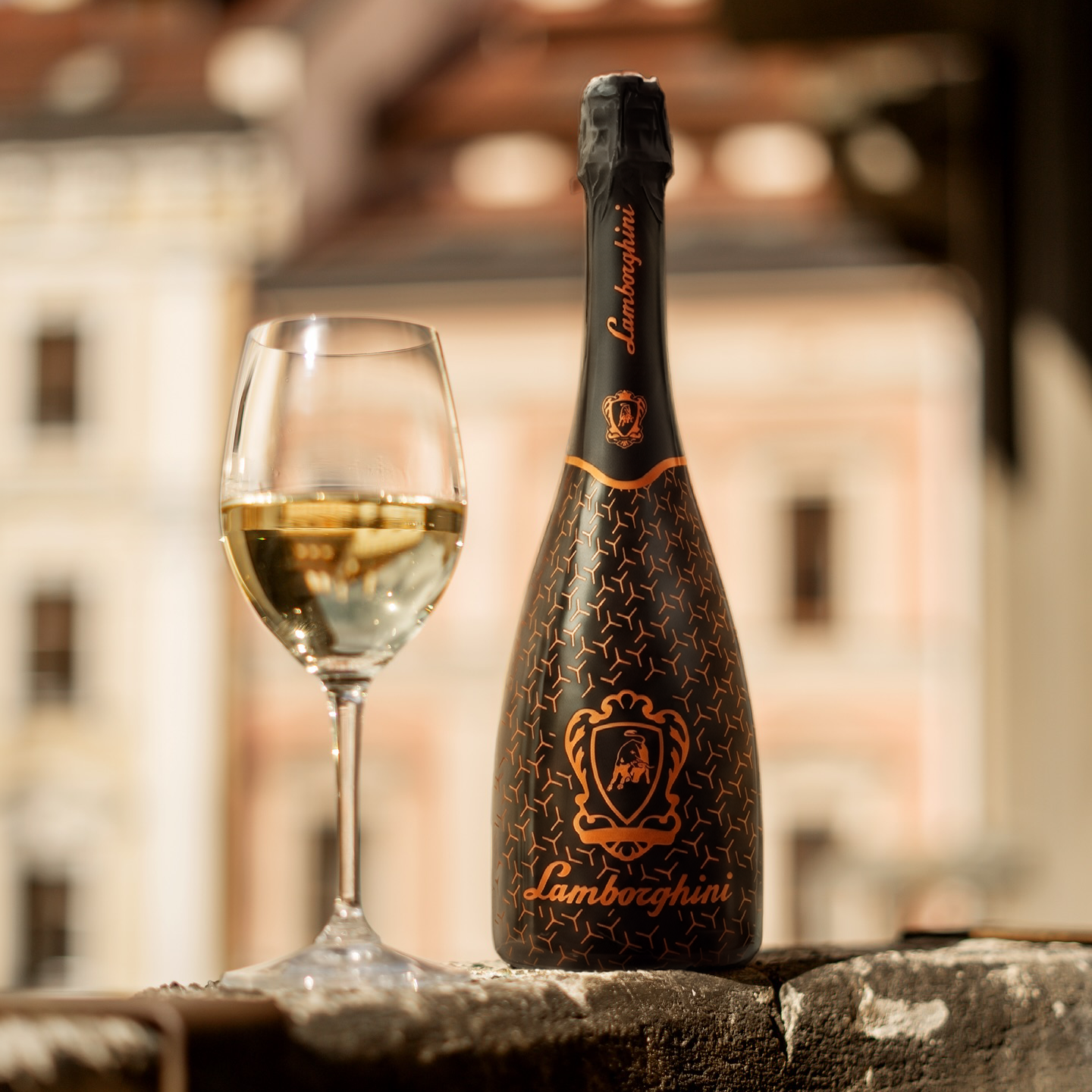 A bottle of Lamborghini sparkling wine with a distinctive bronze/orange graphic pattern and a glass on a stone ledge, with a blurred historic European building in the sunny background.