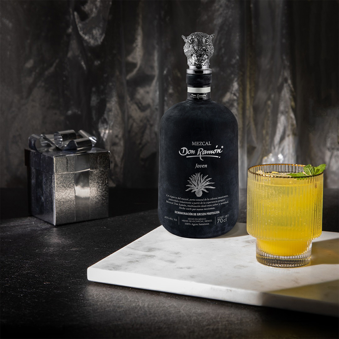 A black bottle of Mezcal Don Ramón Salmiana Joven standing on a marble board. Next to it is a ribbed glass with a yellow cocktail and mint, and an elegant silver gift box in the background.
