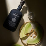 A black Mezcal Don Ramón Espadín Joven bottle lying on a light counter, next to a ceramic plate with pieces of fresh green melon and a small tasting glass in the sunlight.