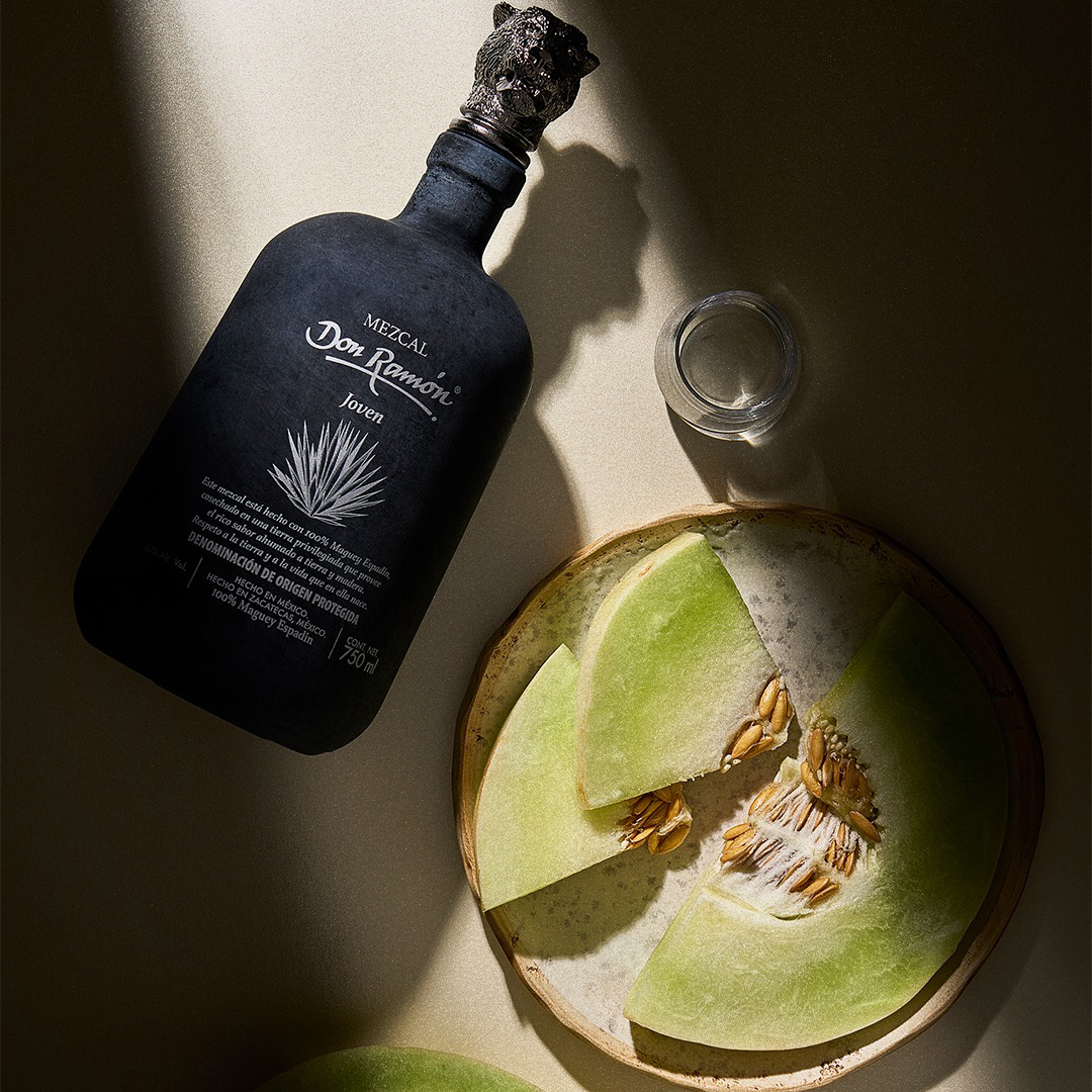 A black Mezcal Don Ramón Espadín Joven bottle lying on a light counter, next to a ceramic plate with pieces of fresh green melon and a small tasting glass in the sunlight.