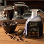 Butelka likieru kawowego Patrón XO Cafe z charakterystycznym korkiem i fioletową wstążką, ustawiona na drewnianym stole obok miseczki z ziarnami kawy i dwóch przezroczystych filiżanek z czarną kawą.