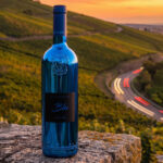 A blue metallic bottle of Lamborghini Luxe Blu wine standing on a stone wall against the backdrop of a vast vineyard and a winding road with car light trails at sunset.
