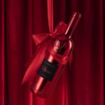 A glossy red bottle of Lamborghini Luxe Rosso wine suspended in the air, tied with a large red ribbon and bow, against a red velvet curtain background.