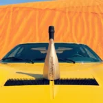 A gold bottle of Lamborghini Icona Oro wine standing majestically on the hood of a classic yellow Lamborghini sports car. Vast, sun-drenched sandy dunes are visible in the background.