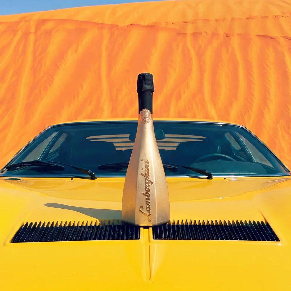 A gold bottle of Lamborghini Icona Oro wine standing majestically on the hood of a classic yellow Lamborghini sports car. Vast, sun-drenched sandy dunes are visible in the background.