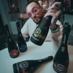A man in pink glasses sits at a table holding a black bottle of Lamborghini DJ Luminous wine with yellow illumination. Other bottles from the Luminous series, glowing blue, pink, and yellow, are on the table.
