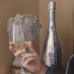 A silver bottle of Lamborghini Extra Dry wine standing next to a classical stone bust of a Greek or Roman sculpture. In the blurred foreground, a man's hand holds a glass of white wine.