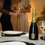 A bottle of Lamborghini Metodo Classico Brut standing on an elegantly set dining table. In the blurred background, two women holding wine glasses during a party.