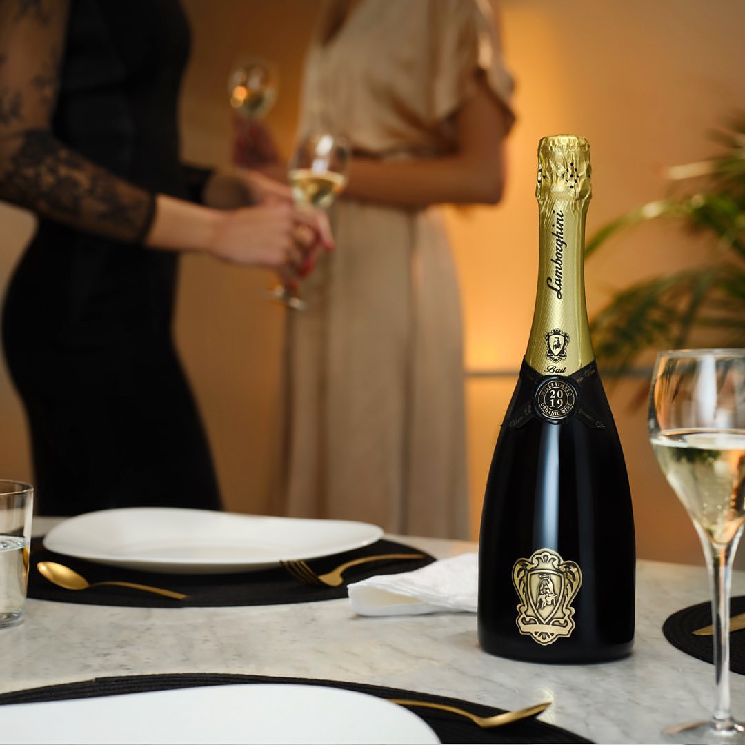 A bottle of Lamborghini Metodo Classico Brut standing on an elegantly set dining table. In the blurred background, two women holding wine glasses during a party.