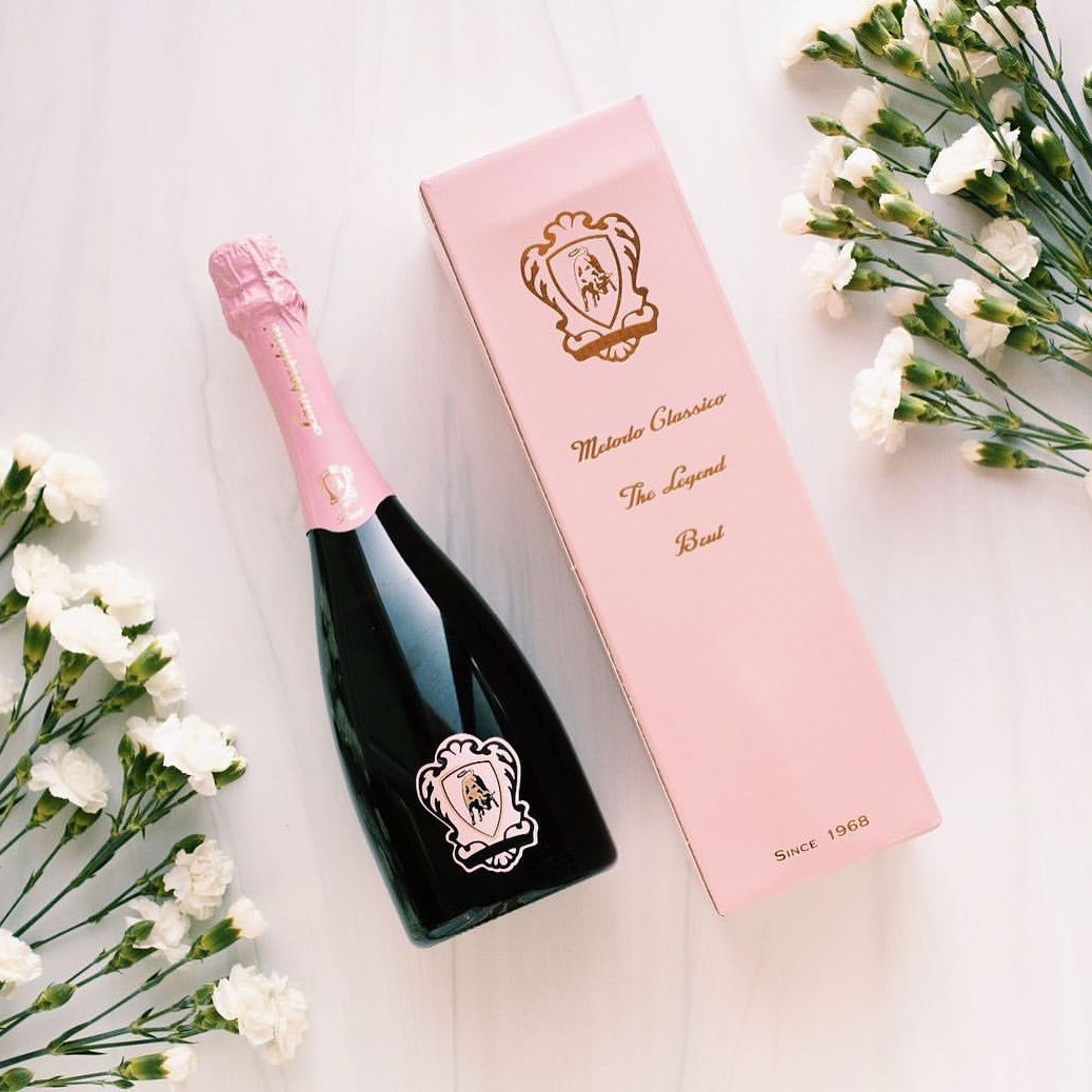 Lamborghini Metodo Classico Rosé sparkling wine lying on a light marble background next to a slim pink gift box and white carnations