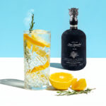 A bottle of Mezcal Don Ramón Salmiana Joven placed next to a tall crystal glass with a refreshing drink full of ice, orange slices, and a smoking sprig of rosemary. Light blue background.