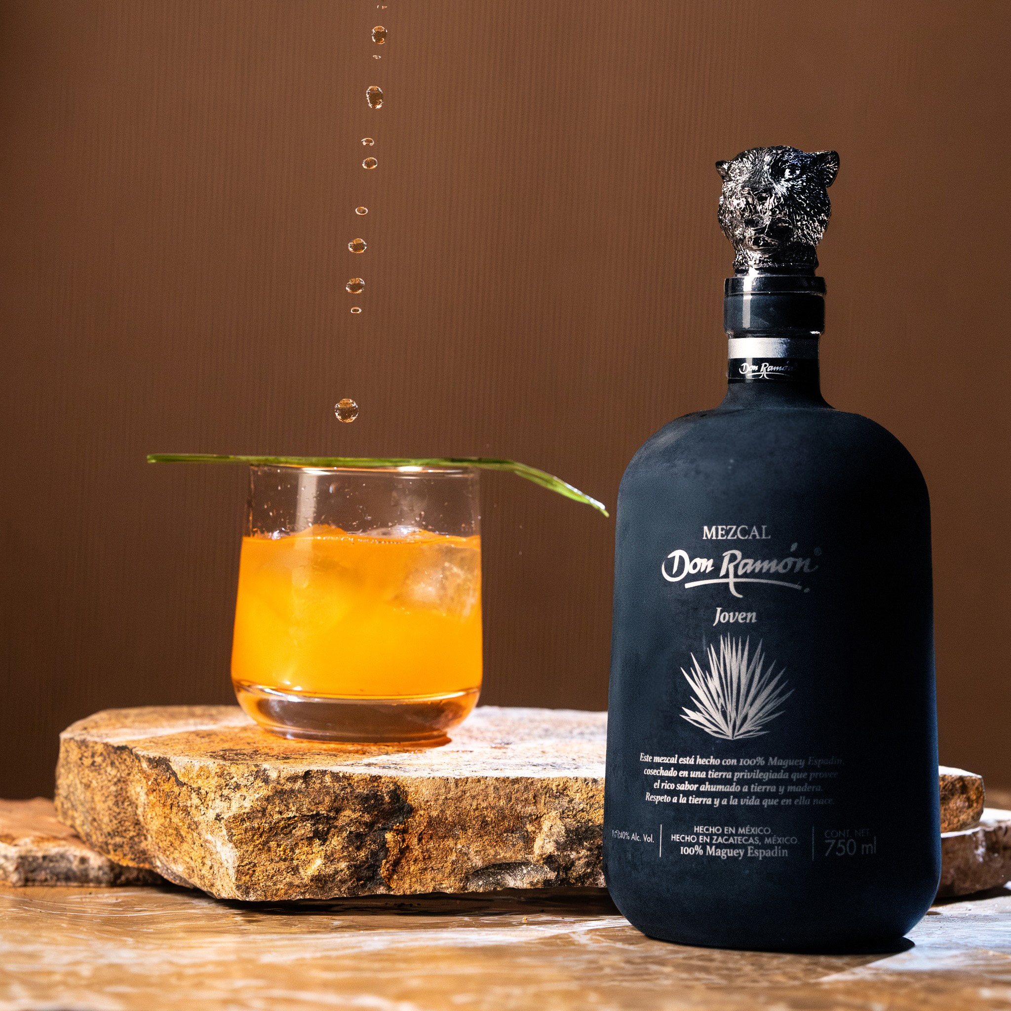 A bottle of Mezcal Don Ramón standing on a raw stone. Next to it is a glass with an orange cocktail and ice, decorated with a green leaf, with drops of liquid hovering in the air above it.