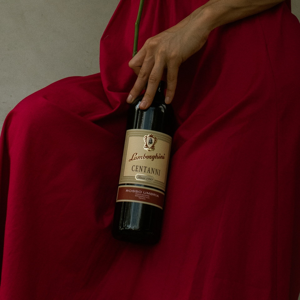 A woman's hand in an elegant red dress holding a bottle of Lamborghini Centanni Rosso Umbria red wine resting against the dress material.