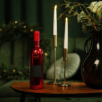 A red, glossy Lamborghini Luxe Rosso bottle standing on a wooden table accompanied by two lit candles and a dark vase with flowers against a green sofa background.