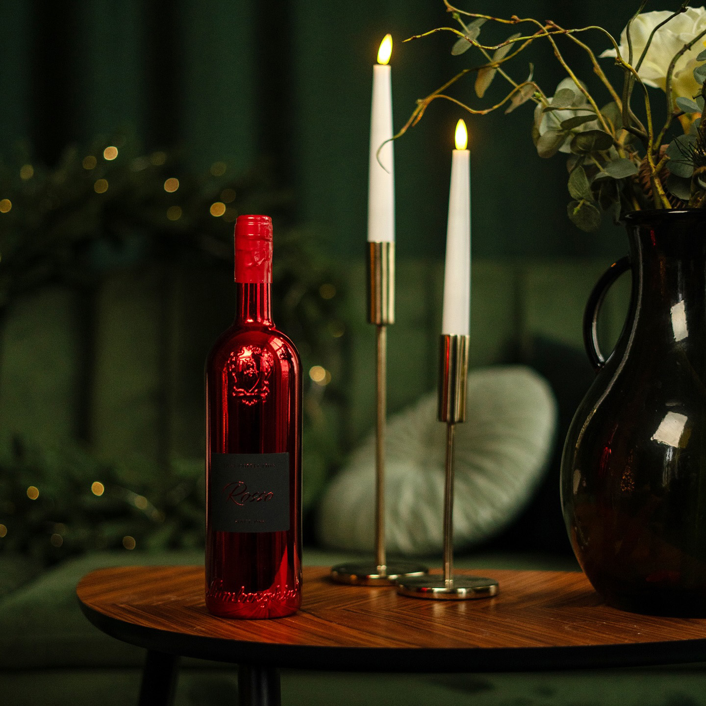 A red, glossy Lamborghini Luxe Rosso bottle standing on a wooden table accompanied by two lit candles and a dark vase with flowers against a green sofa background.