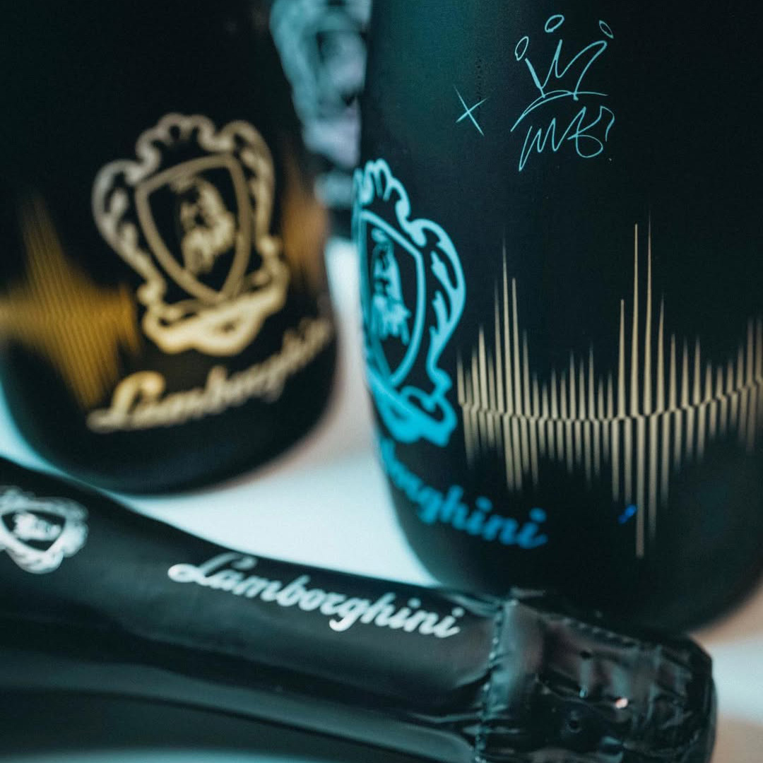 Close-up of a black Lamborghini DJ Luminous bottle with a blue-illuminated logo and a graffiti-style autograph on the top of the bottle. A second bottle lies nearby, partially visible